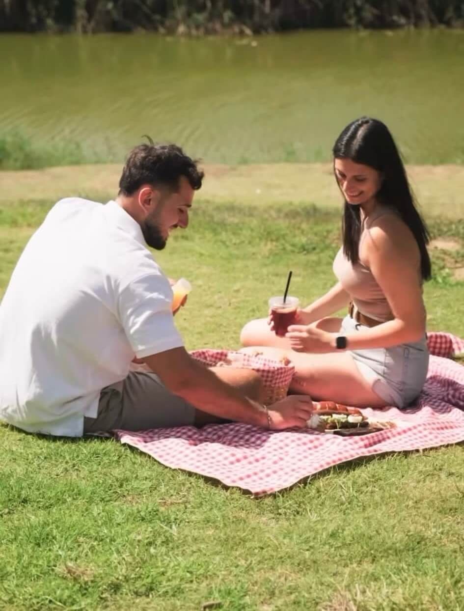 זוג יושב על מחצלת פיקניק על הדשא ליד נחל לכיש, עם קפה ומגש אוכל מהעגלה